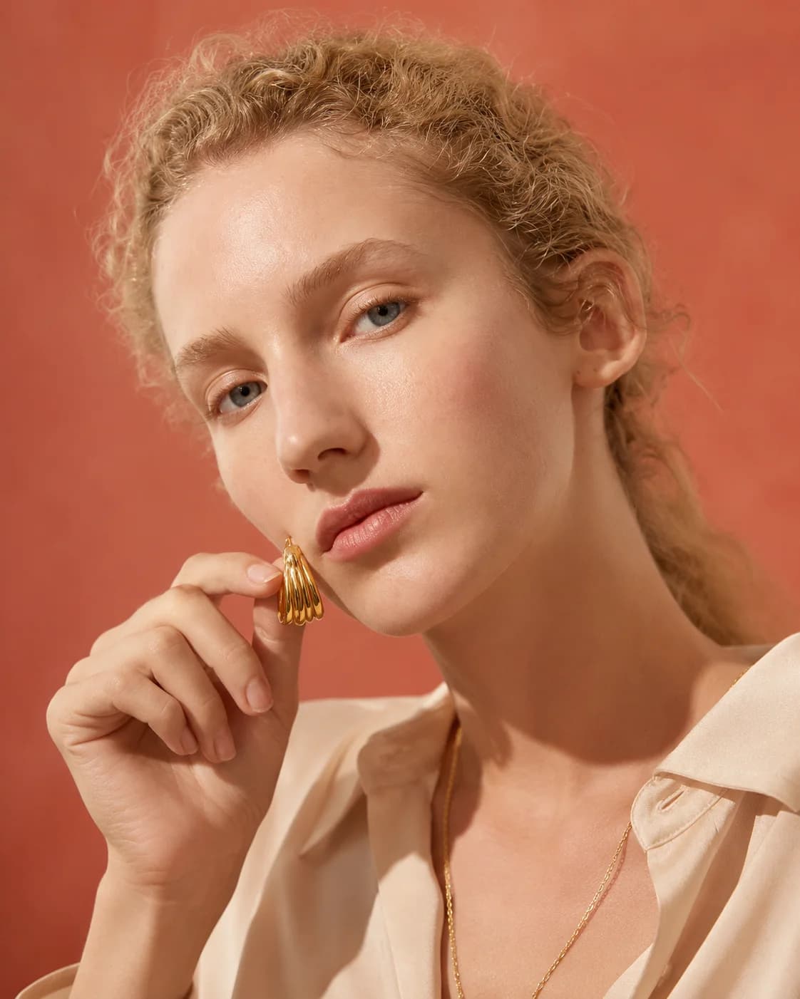 A woman holding a yellow gold earring against her face, on a warm terracotta ground