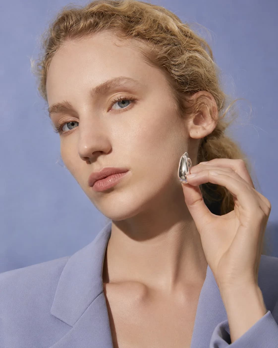 The same woman holding a silver earring against her face, on a cool blue ground